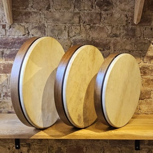 May include: Three wooden frame drums of different sizes, with natural colored drumheads and black trim. The drums are arranged in a row from largest to smallest.