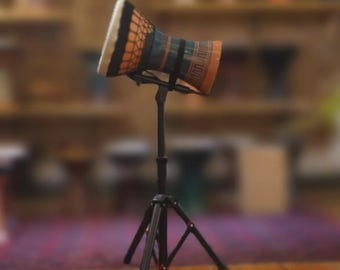 May include: A small, hand-held drum with a white top, orange and blue body, and black accents. The drum is mounted on a black tripod stand, set against a blurred background of a room with wooden shelves and a patterned rug.
