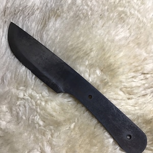 May include: A black, unfinished knife blade with a rounded tip and a handle with two holes. The knife is lying on a white, fluffy surface.