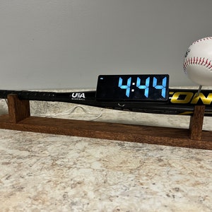 May include: A black baseball bat with a digital clock displaying 4:44 on top of it. The bat is on a wooden stand with a white baseball on top of the bat.