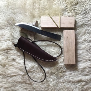 May include: A handmade knife with a stainless steel blade and a dark brown leather sheath. The sheath is stitched with white thread. Also included are wooden blocks and brass pins, likely for handle construction.