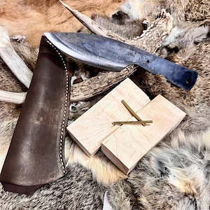 May include: A dark brown leather sheath with a stitched edge, a black metal knife with a curved blade, and two gold metal rods on a light brown wooden block.