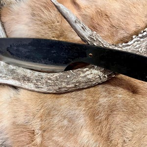 Könnte beinhalten: Ein schwarzes, handgeschmiedetes Messer mit einem Holzgriff. Das Messer liegt auf einem Stück Tierfell.