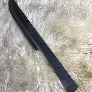 May include: A black metal knife blade with a rounded tip and a handle with four holes. The blade is lying on a white fluffy surface.