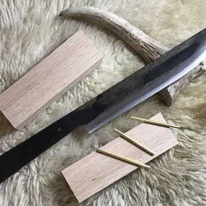 May include: A long, black, metal knife with a wooden handle. The knife is lying on a white, fluffy surface. There are two pieces of wood and four brass rods on the surface.
