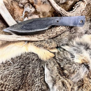 Puede incluir: Un cuchillo negro forjado a mano con mango de cuero, descansando sobre una pila de pieles de animales. El cuchillo tiene una punta afilada y una hoja ligeramente curvada.