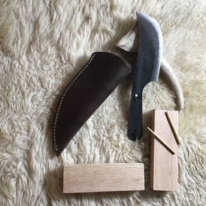 May include: A brown leather sheath with a curved blade knife. The knife has a wooden handle and a metal blade. The knife is on a white furry surface with two wooden blocks.