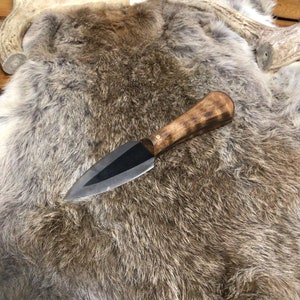 Peut inclure: Un couteau à manche noir avec un manche en bois brun foncé et une lame pointue et tranchante. Le couteau est posé sur une peau de fourrure brune et grise.