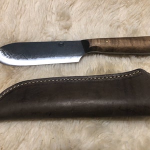 May include: A brown leather sheath with a stitched edge, holding a  handmade knife with a wooden handle. The knife has a  curved blade and a  silver metal accent.