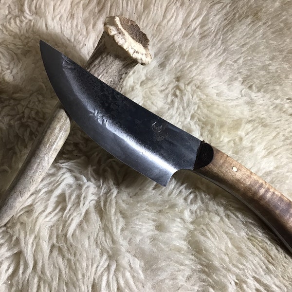 Puede incluir: Un cuchillo negro forjado a mano con mango de madera. La hoja tiene una ligera curva y una punta redondeada. El mango es de madera con una mancha marrón oscura. El cuchillo está apoyado sobre una superficie blanca y esponjosa.