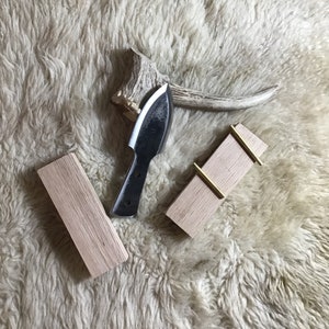 May include: Two wooden blocks with brass accents, one with a small knife with a black handle and a curved blade. The blocks and knife are on a white fluffy surface.