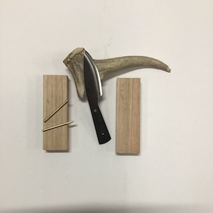 May include: Three wooden blocks with a knife and an antler on a white background. The blocks are made of light wood and have a natural grain. The knife is black with a silver blade. The antler is brown and has a natural curve. Two gold metal rods are attached to the left block.