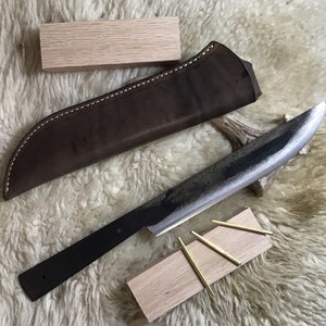 May include: A black knife with a leather sheath and wooden handle. The knife is on a white fluffy surface with a wooden block and brass pins.