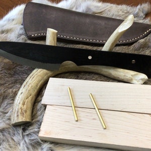 May include: A black knife with a wooden handle and a leather sheath. The knife is displayed on a piece of antler. Two brass rods are on a wooden block.