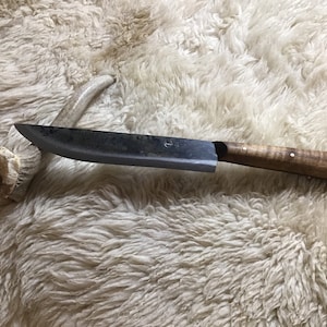May include: A long, curved, black knife with a wooden handle. The handle is light brown with darker brown accents. The knife is lying on a white, fluffy surface.