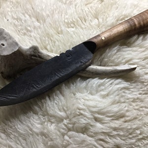 Könnte beinhalten: Ein schwarzes, handgeschmiedetes Messer mit einem Holzgriff. Der Griff hat ein Maserungsmuster und einen Messingpin. Das Messer liegt auf einer weißen, flauschigen Oberfläche.