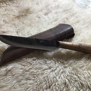 May include: A curved, black-handled knife with a brown leather sheath. The knife has a slightly curved blade and a wooden handle with a light brown finish.