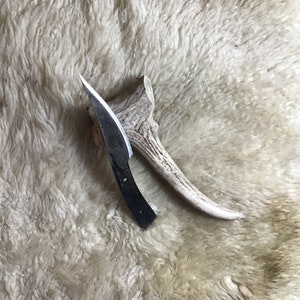 May include: A handmade knife with a curved blade and a dark handle rests beside a piece of antler. The knife has a sharp, pointed tip and a dark, textured blade. The antler is a light brown color.
