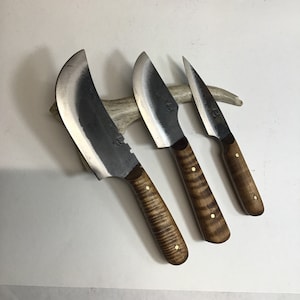 May include: Three handmade knives with wooden handles. The largest knife has a curved blade and the two smaller knives have straight blades. All three knives have brass accents on the handles.