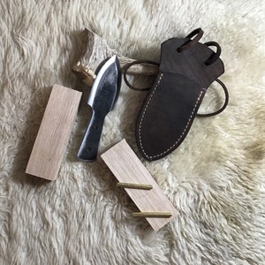 May include: A small, black, hand-forged knife with a leather sheath and two wooden blocks. The knife has a curved blade and a handle made of wood. The sheath is brown leather with a strap and buckle. The wooden blocks are light brown and have a smooth surface.
