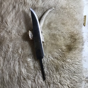May include: A long, thin, silver knife blade with a curved edge. The blade is resting on a piece of white fur.
