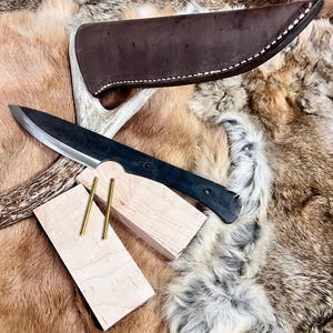 May include: A black knife with a wooden handle and a leather sheath. The knife is resting on a piece of wood with two brass pins. The wood is on a brown and white fur.