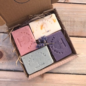May include: A cardboard gift box containing four Cherry Tree Naturals soaps. The soaps are rectangular and in shades of pink, white, purple, and green. Each soap is stamped with the brand name and a tree logo. A sprig of lavender is on top.