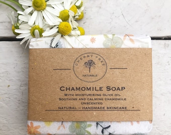 May include: A bar of chamomile soap wrapped in floral patterned paper. The soap is labeled "Cherry Tree Naturals Chamomile Soap with Moisturizing Olive Oil, Soothing and Calming Chamomile, Unscented, Natural Handmade Skincare."