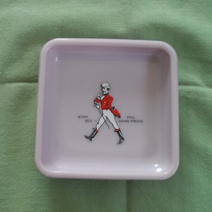 May include: A white square ceramic ashtray with a red and white logo of a man in a red jacket and white pants walking with a cane. The logo says "Born 1820 - Still Going Strong".