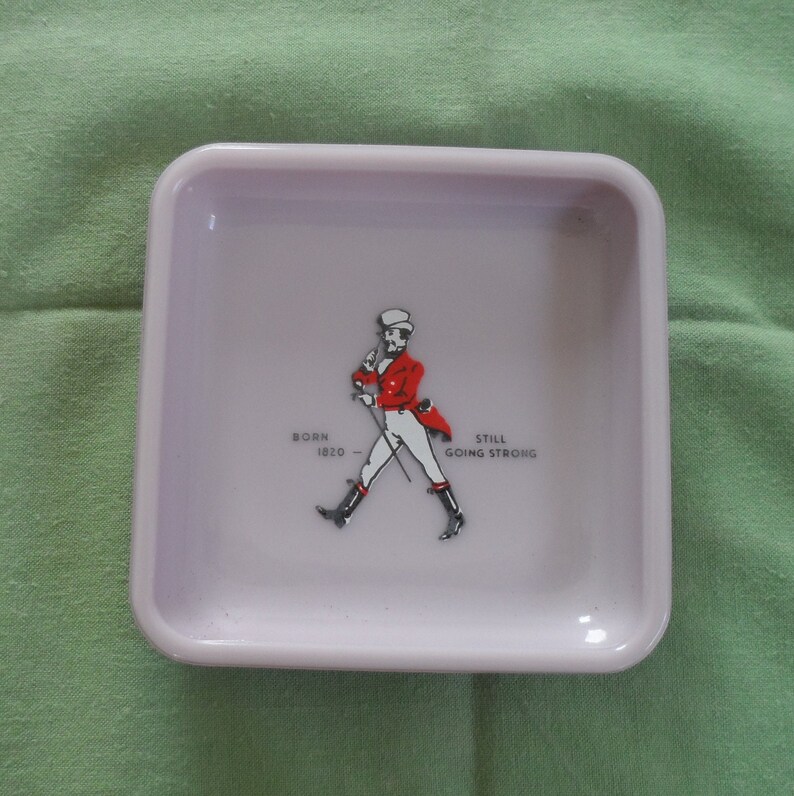 May include: A white square ceramic ashtray with a red and white logo of a man in a red jacket and white pants walking with a cane. The logo says "Born 1820 - Still Going Strong".