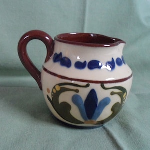 May include: A small, white ceramic creamer with a brown handle. The creamer has a blue and green floral design with yellow accents.
