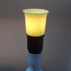 May include: A small lamp with a pale yellow shade and a light green rim. The shade sits on a black base, connected to a light blue, ornate base. The lamp emits a soft, warm glow, creating a gentle ambiance.