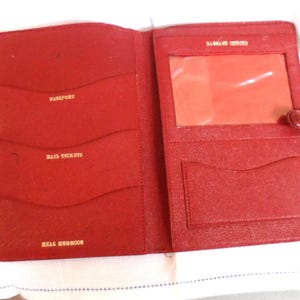 May include: Open red leather travel wallet with multiple pockets for passport, rail tickets, and other documents. The wallet has a clear window for identification and a snap closure. The words "Passport", "Rail Tickets", and "Real Morocco" are printed in gold.