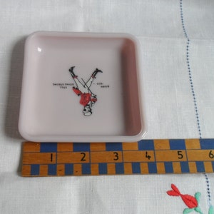 May include: A small, square, pink, ceramic ashtray with a graphic of a man in a red jacket and white pants standing on his hands. The text "BORN - 026. STILL GOING STRONG" is printed on the ashtray.