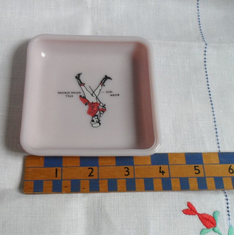 May include: A small, square, pink, ceramic ashtray with a graphic of a man in a red jacket and white pants standing on his hands. The text "BORN - 026. STILL GOING STRONG" is printed on the ashtray.
