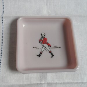 May include: A pink square ceramic ashtray with a black and white image of a man in a red jacket and white pants walking with a cane. The text "BORN 1620 - STILL GOING STRONG" is printed below the image.