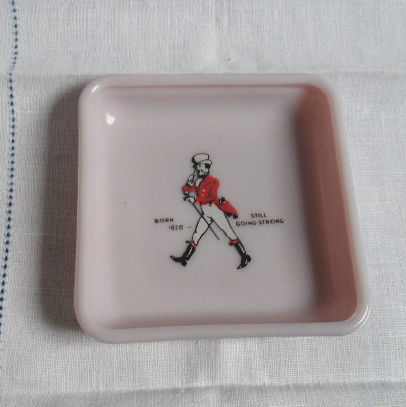 May include: A pink square ceramic ashtray with a black and white image of a man in a red jacket and white pants walking with a cane. The text "BORN 1620 - STILL GOING STRONG" is printed below the image.