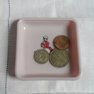 May include: A pink ceramic dish with three British coins. The coins are a half crown, a sixpence, and a shilling. The half crown has the image of Queen Elizabeth II on it. The sixpence has the image of a ship on it. The shilling has the image of a lion on it.