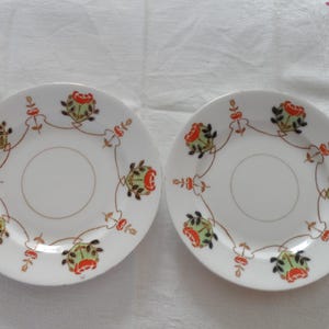 May include: Two white saucers with a floral design. Each saucer features a central circle and is decorated with a repeating pattern of red and green flowers, connected by a gold-colored line. The saucers are set against a white background.