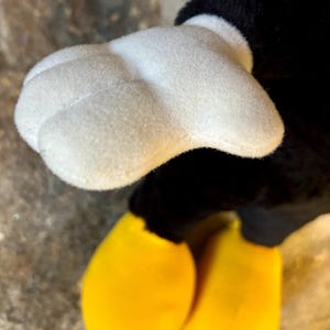 以下が含まれることがあります： ぬいぐるみのおもちゃの白い手袋の手と黄色の足のクローズアップ。おもちゃの黒い体と頭が背景に見えます。ぬいぐるみのおもちゃは、古典的な漫画のキャラクターです。