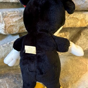 以下が含まれることがあります： 黒いミッキーマウスのぬいぐるみ。白い手袋と黄色の足があります。おもちゃにはタグが付いています。背景は石壁です。後ろから見たところ。