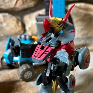 Puede incluir: Un juguete robot articulado y colorido con una combinación de colores rojo, dorado y gris. El juguete tiene un diseño detallado con múltiples articulaciones y una apariencia estilizada. Otro juguete es visible en el fondo.