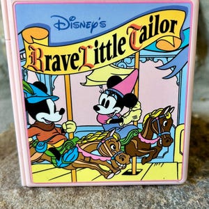 May include: A pink, rectangular Disney's Brave Little Tailor compact featuring Mickey and Minnie Mouse on a carousel. The artwork includes a yellow banner with the title, set against a light blue and pink background. The compact has a glossy finish.