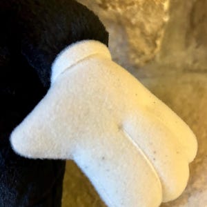 以下が含まれることがあります： 黒いぬいぐるみのアームに付いた、3本指と親指のある白いぬいぐるみ手袋のクローズアップ。手袋は柔らかく、質感のある外観をしています。背景はぼやけた、薄茶色の石壁です。