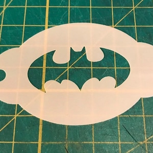Puede incluir: Plantilla de plástico blanco con un logotipo de Batman recortado. La plantilla tiene forma ovalada.