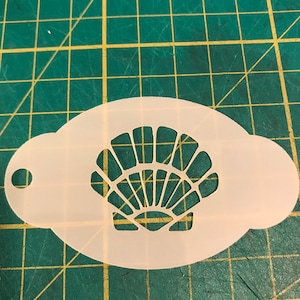 May include: White plastic stencil featuring a seashell design. The stencil has a handle on the side.
