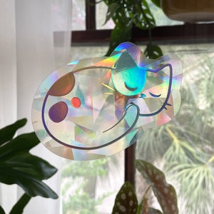 May include: A holographic sticker of a sleeping cat with brown spots and a blue ear. The sticker has a rainbow iridescent finish.