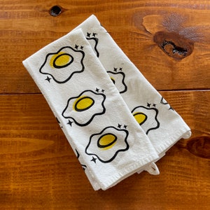 May include: Two white kitchen towels with a black and yellow fried egg design. The eggs are outlined in black and have a yellow yolk.