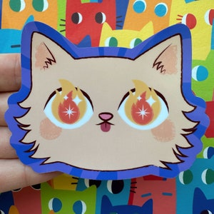 May include: A sticker of a cartoon cat with blue fur, a light brown face, and large orange eyes with flames and sparkles. The cat has a small pink nose and a pink tongue sticking out.