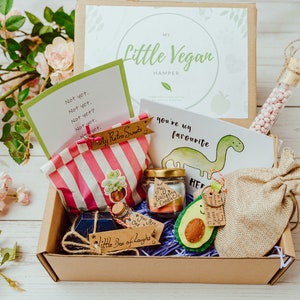 May include: A brown cardboard box filled with vegan treats, including a card that says "You're my favourite" with a dinosaur illustration, a bag of sweets, and a felt avocado. The box is labeled "My Little Vegan Hamper".